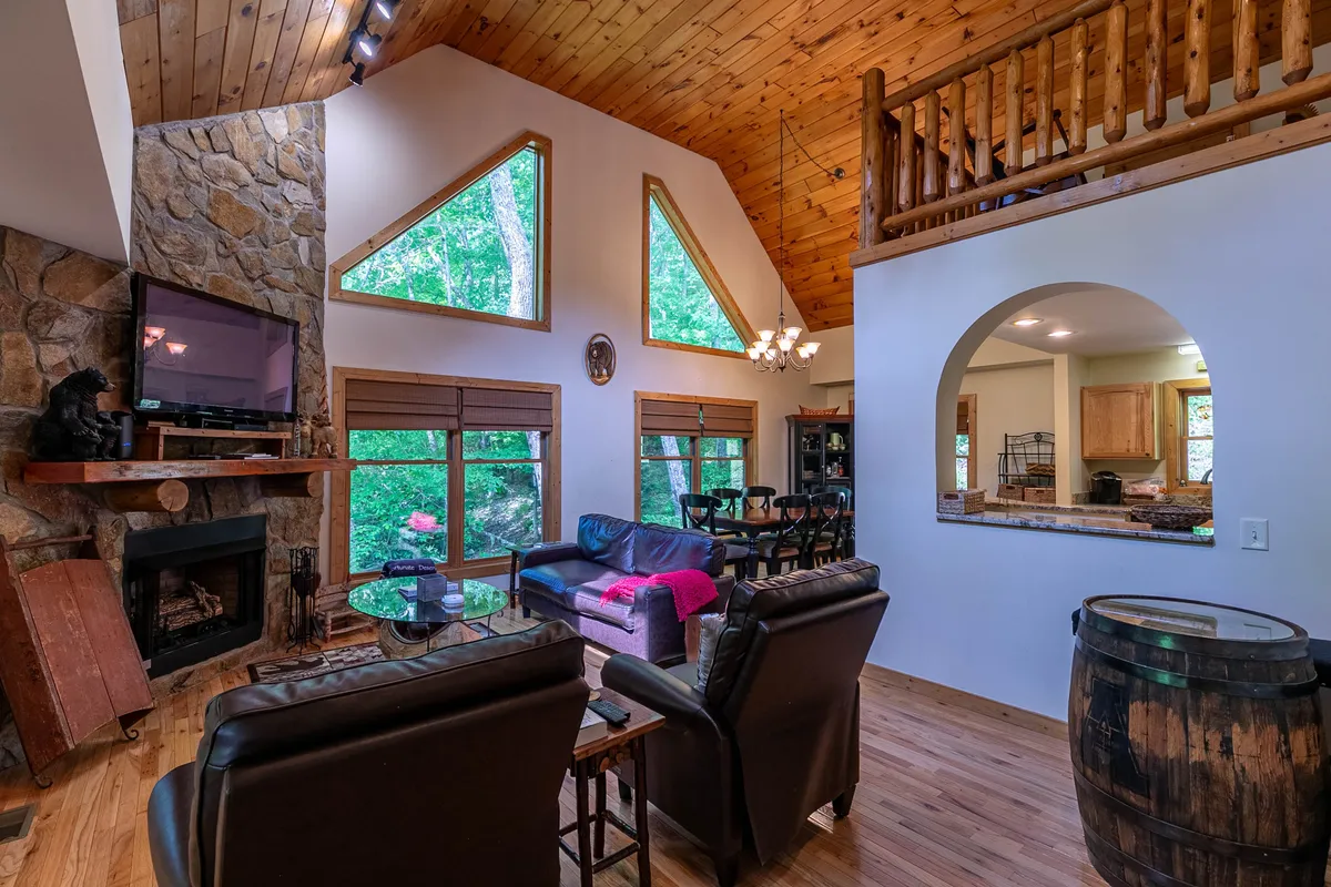 A spacious living area featuring a striking stone fireplace, large windows offering views of the surrounding forest, exposed wooden beams, and a comfortable seating area arranged for socializing or relaxing. The space exudes an inviting atmosphere perfect for family gatherings.