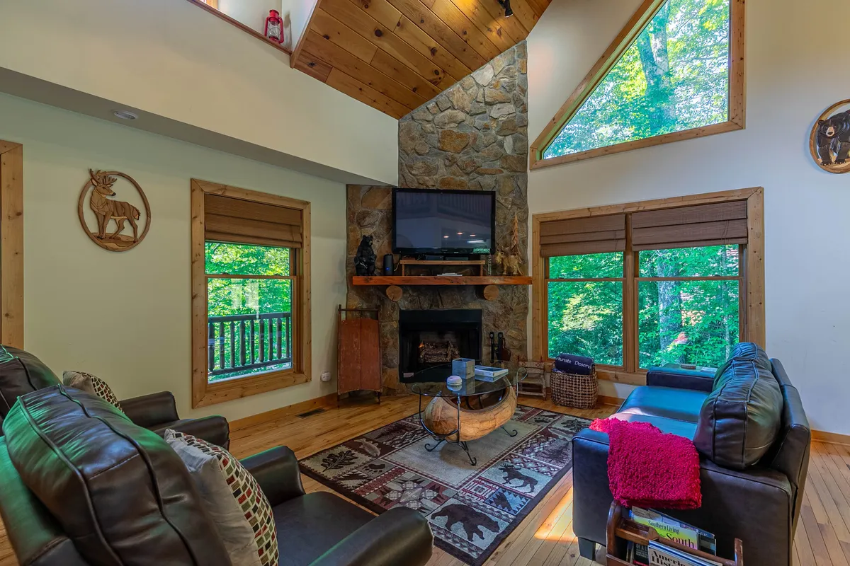 This interior image displays a spacious living room featuring a stone fireplace, large windows that let in abundant natural light, comfortable seating, and decorative wall art. The view through the windows hints at the outdoors, creating a visual connection between the cozy interior and the mountain setting.