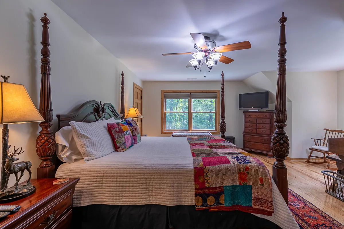 A welcoming bedroom featuring a four-poster bed with a colorful quilt, a large window offering scenic views, a wooden dresser, and a nightstand with a lamp. The window creates a sense of connection to the natural landscape outside, inviting both people and pets to relax and enjoy the surroundings.