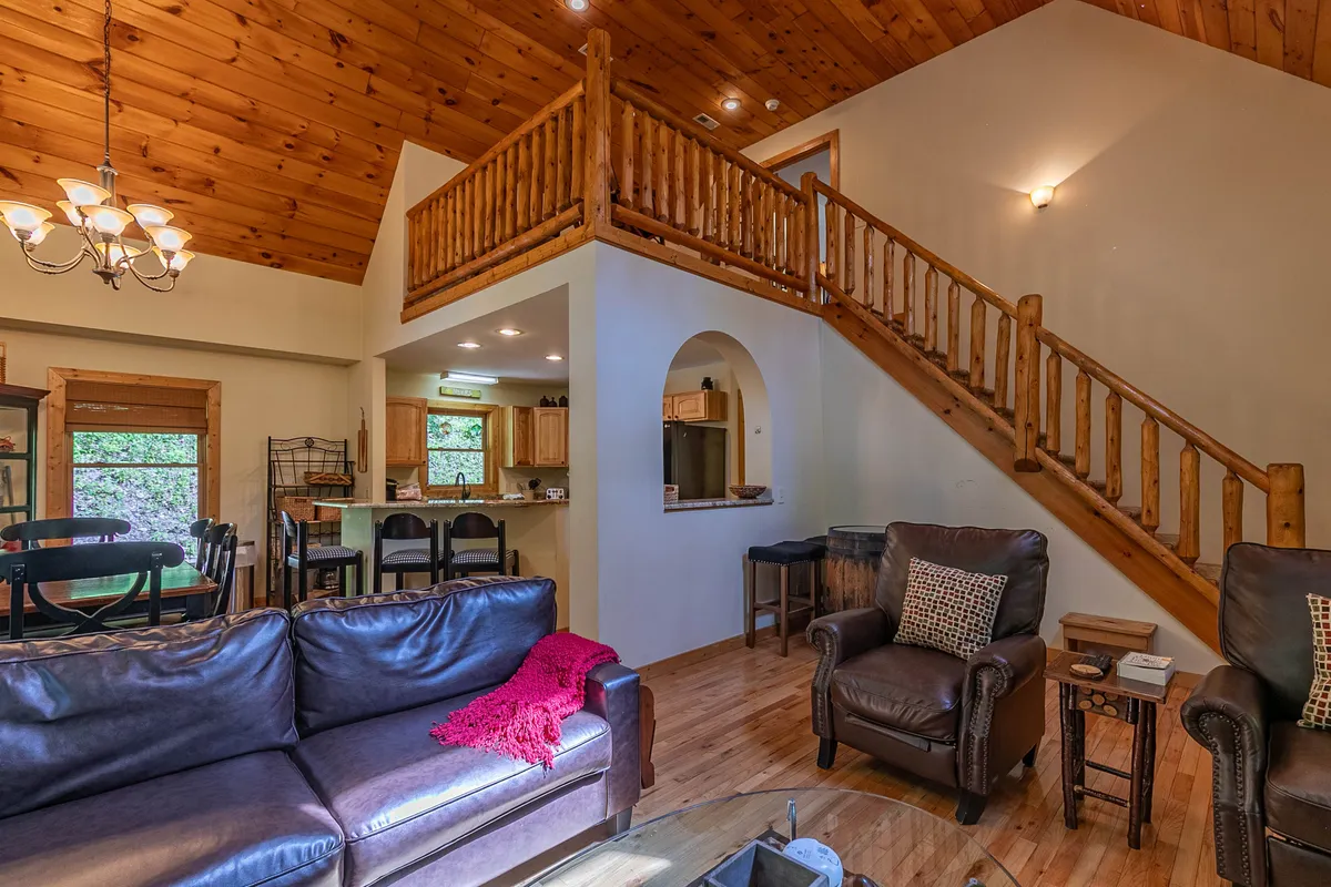 An inviting open-concept living area showcasing a plush sofa, dining table, and visible kitchen area. The wooden ceiling and staircase reinforce the cabin's rustic charm, while large windows provide ample natural light and views of the surrounding landscape.