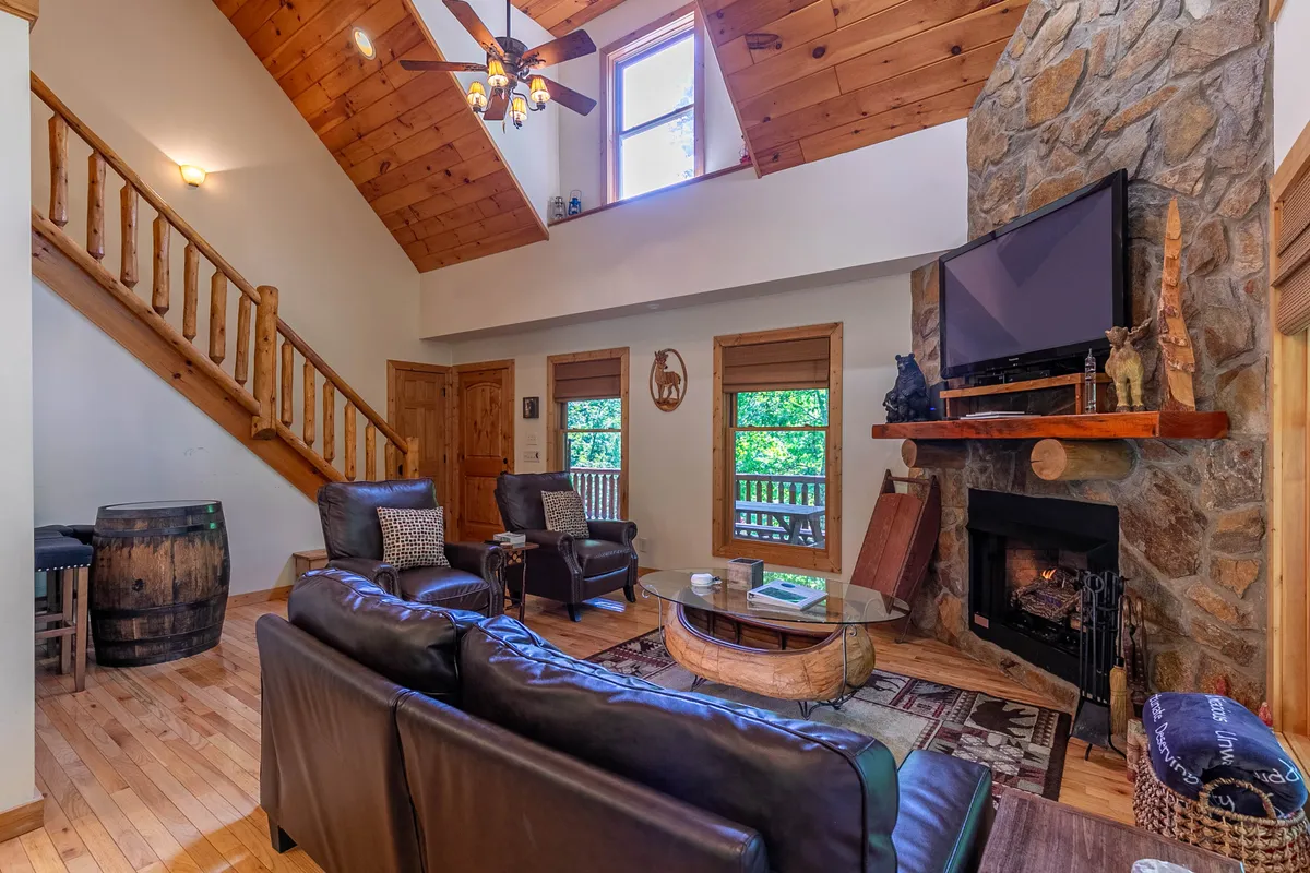 A warm, welcoming living room with a prominent stone fireplace as the focal point. Large windows flood the space with natural light, while a comfortable seating area invites family and friends to gather. The rustic wooden staircase and timber beams add authentic mountain character.