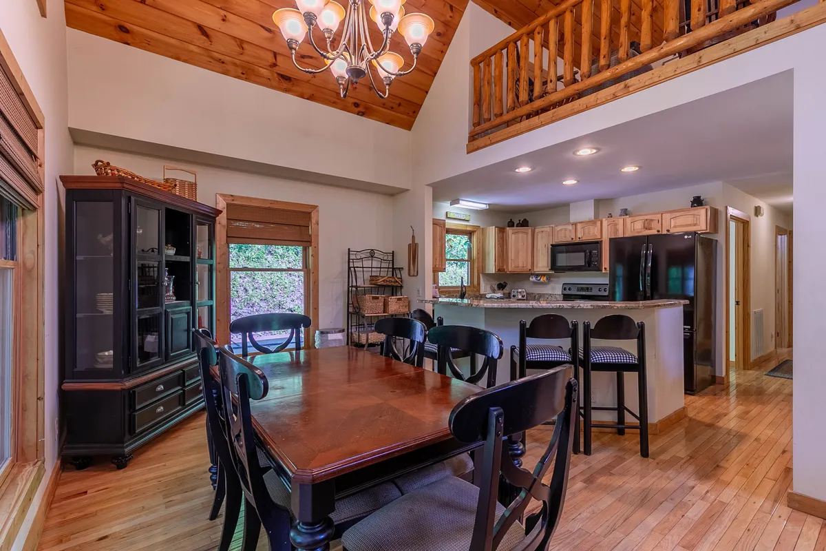 A cozy dining space with a large wooden table under a glowing chandelier, adjacent to a kitchen island and framed by expansive windows that fill the room with light. The arrangement encourages group meals, board games, and lively conversation.