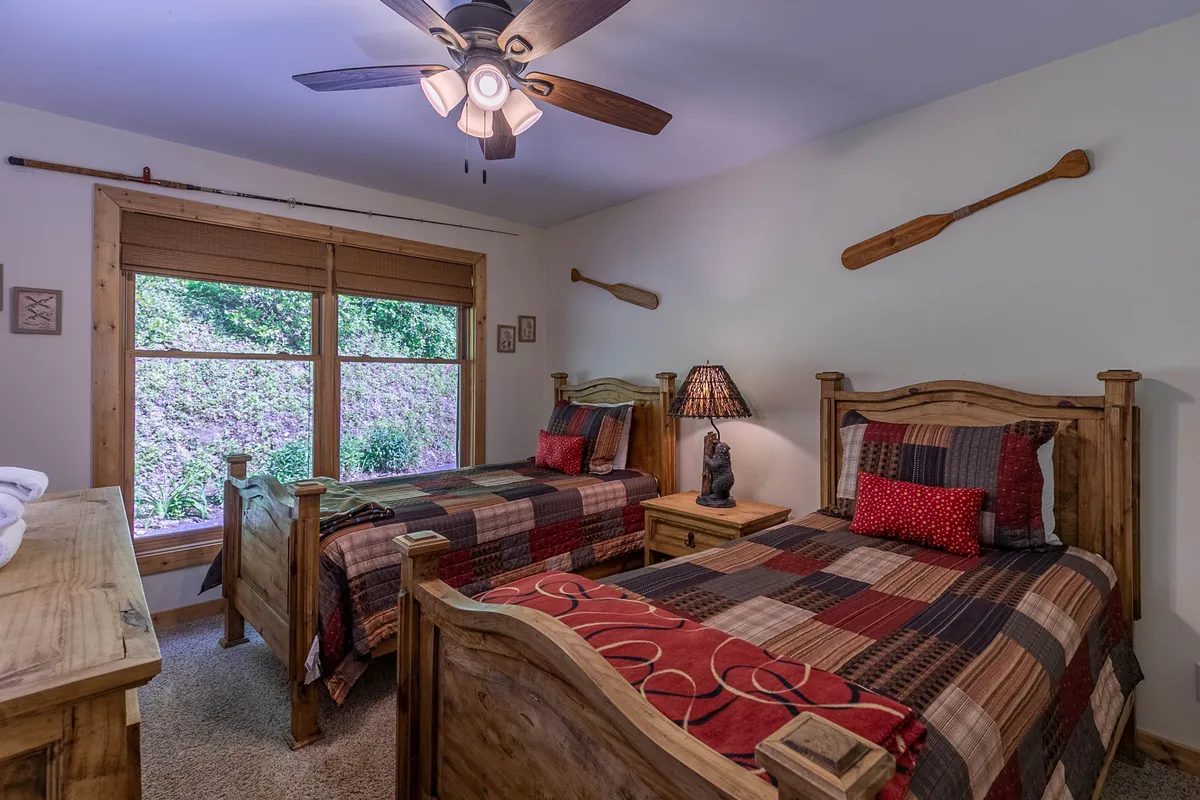 A welcoming twin bedroom with two wooden beds, a table lamp, and playful decorative oars on the wall. A window with an outdoor view invites natural light, making it ideal for kids or friends seeking a cozy retreat.