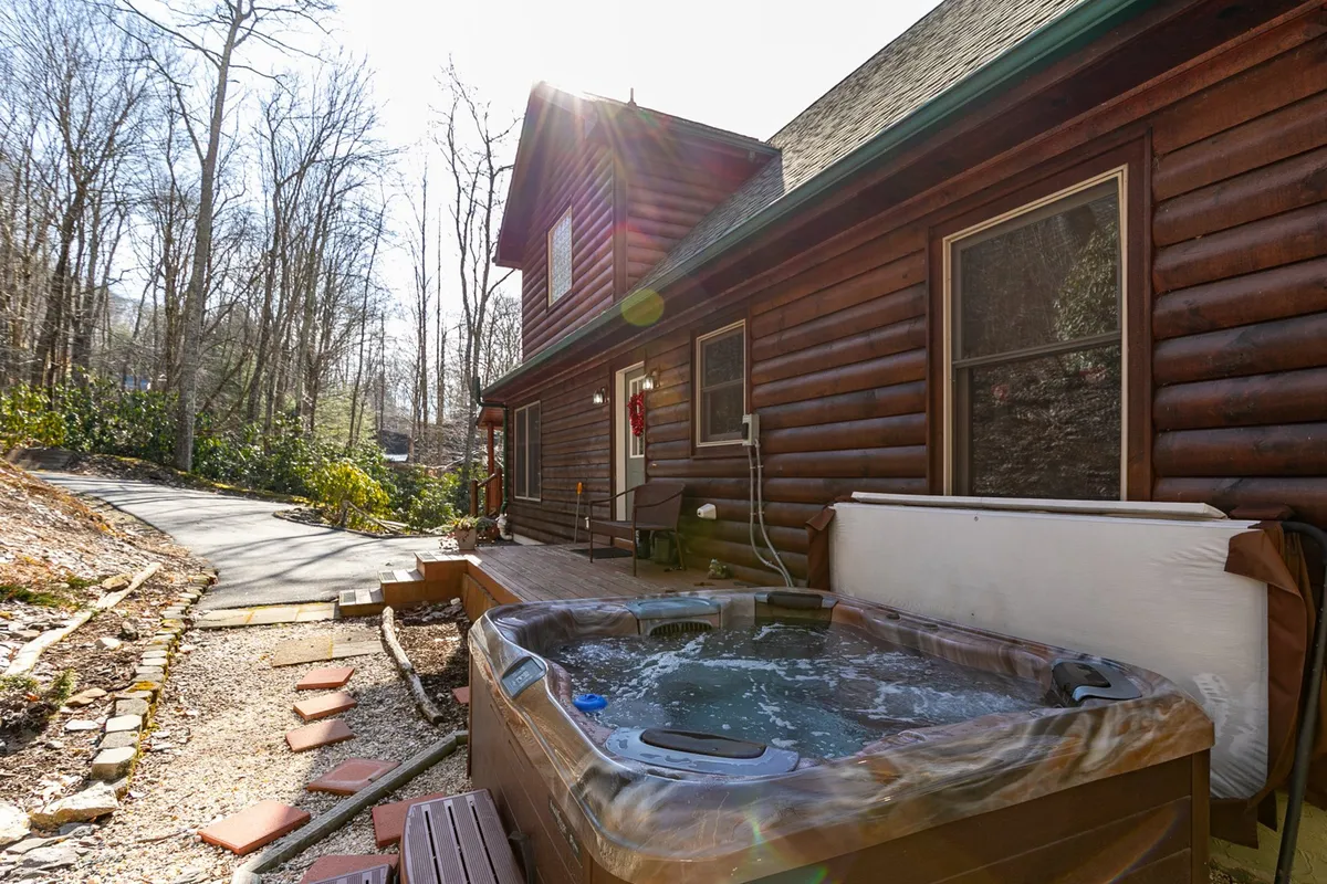 A tranquil shot highlighting the bubbling hot tub nestled beside the log cabin, with a clear view of the surrounding natural landscape and a stone pathway leading to the tub. The scene is perfect for relaxing after outdoor adventures or stargazing in the evening.