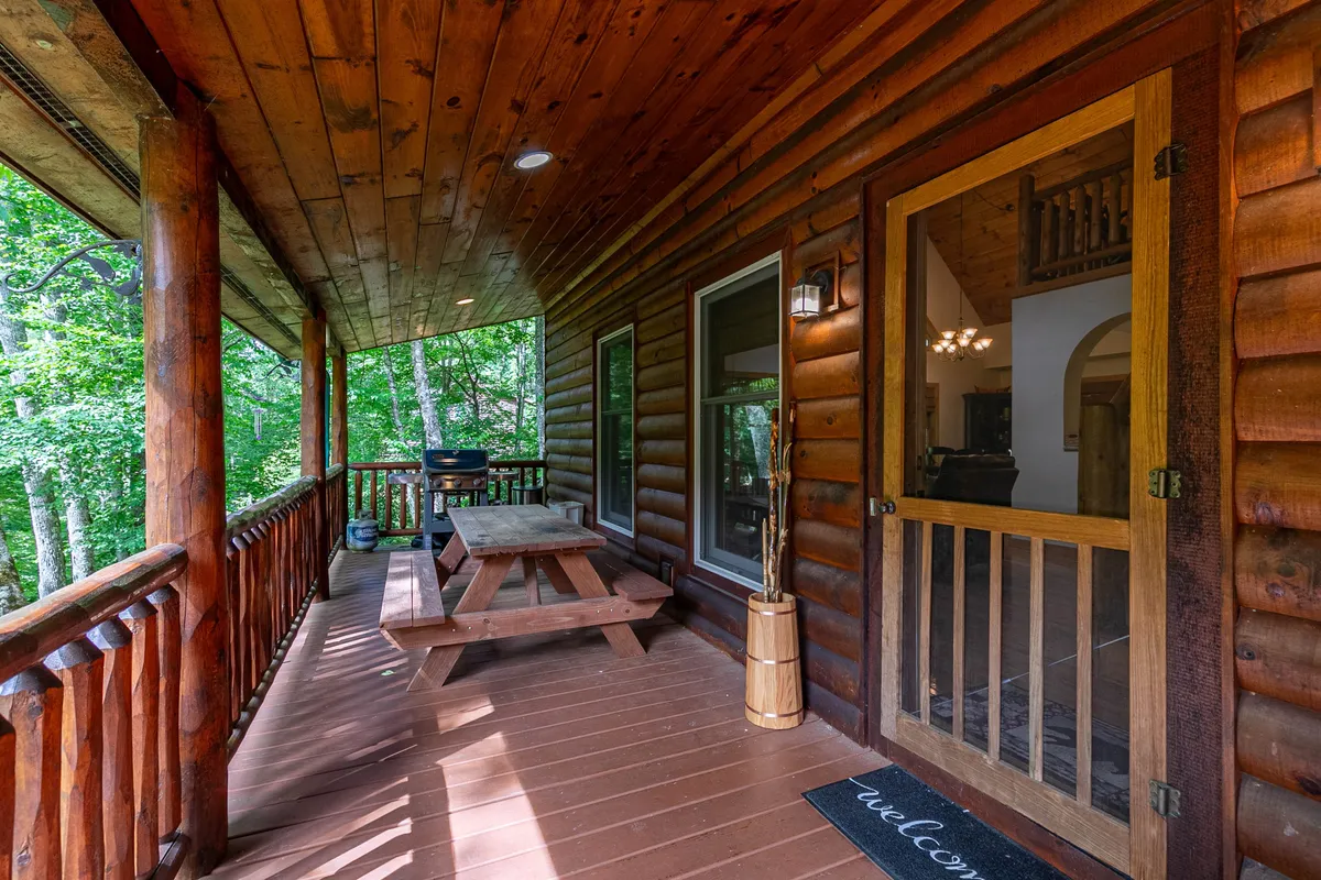 A welcoming outdoor scene featuring a wooden picnic table on an expansive deck, with the log cabin structure and a doorway entrance visible. Surrounded by lush greenery, the setting is ideal for outdoor dining, family gatherings, and enjoying nature.