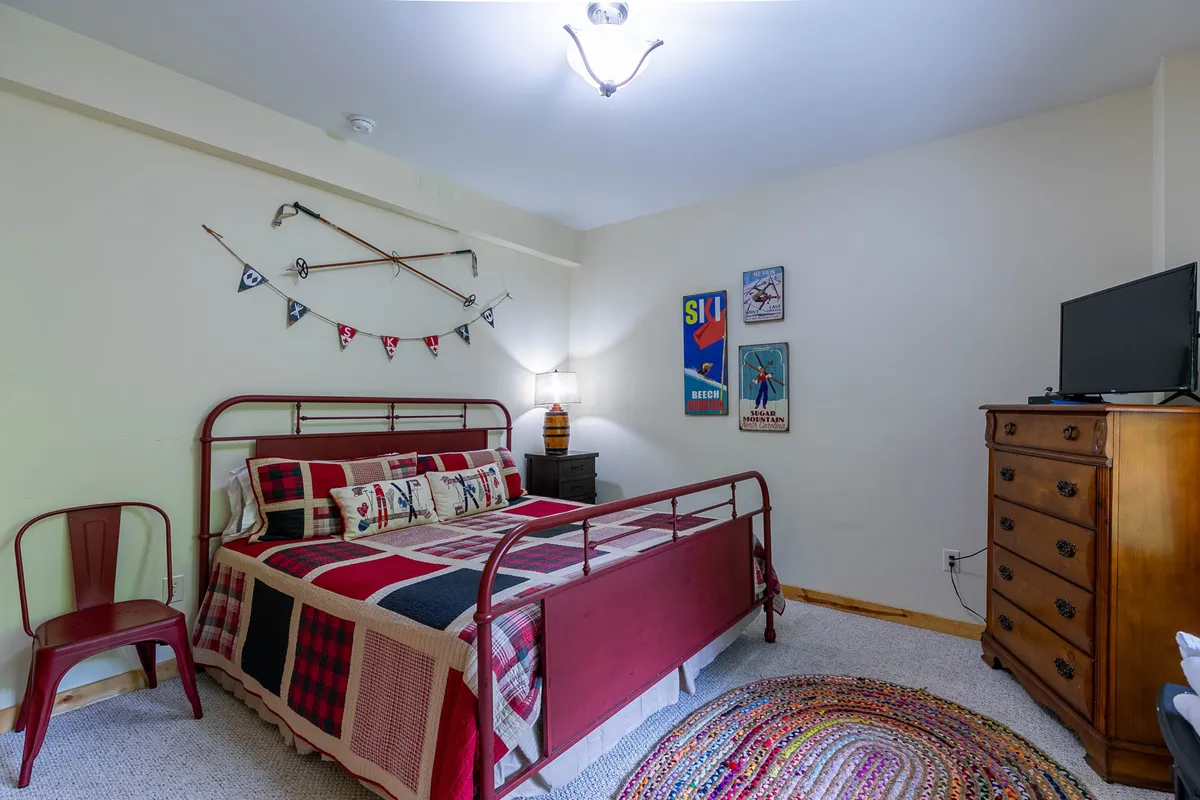 A lower-level bedroom with a bed covered in a colorful quilt, a wooden dresser topped with a TV, decorative wall art, and a cozy rug. The room exudes warmth and offers a private retreat for rest after a lively evening.