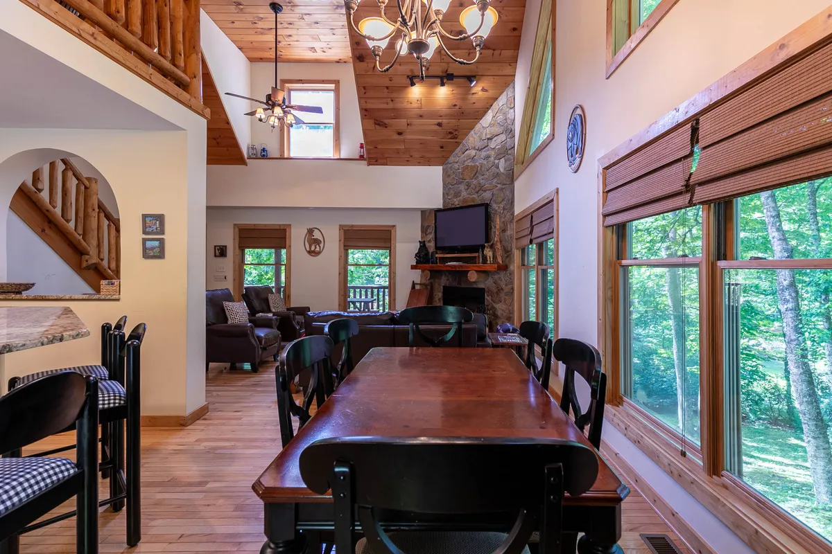 An inviting open-plan area with a large dining table, stone fireplace, wooden ceiling, and comfortable seating—all set against wide windows that bring nature views inside. The space is perfectly suited for group gatherings or relaxed evenings in.